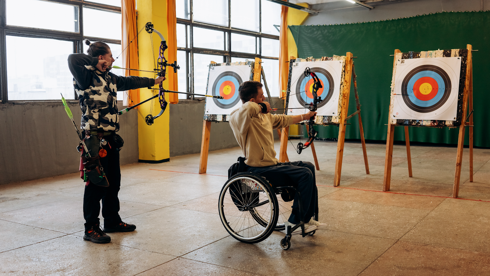 man wearing camo shirt and man in wheelchair aim arrows at targets